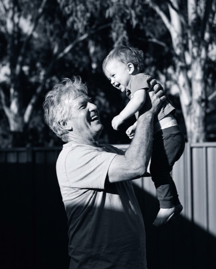 A grandfather lifting his young grandchild up in a backyard, both laughing